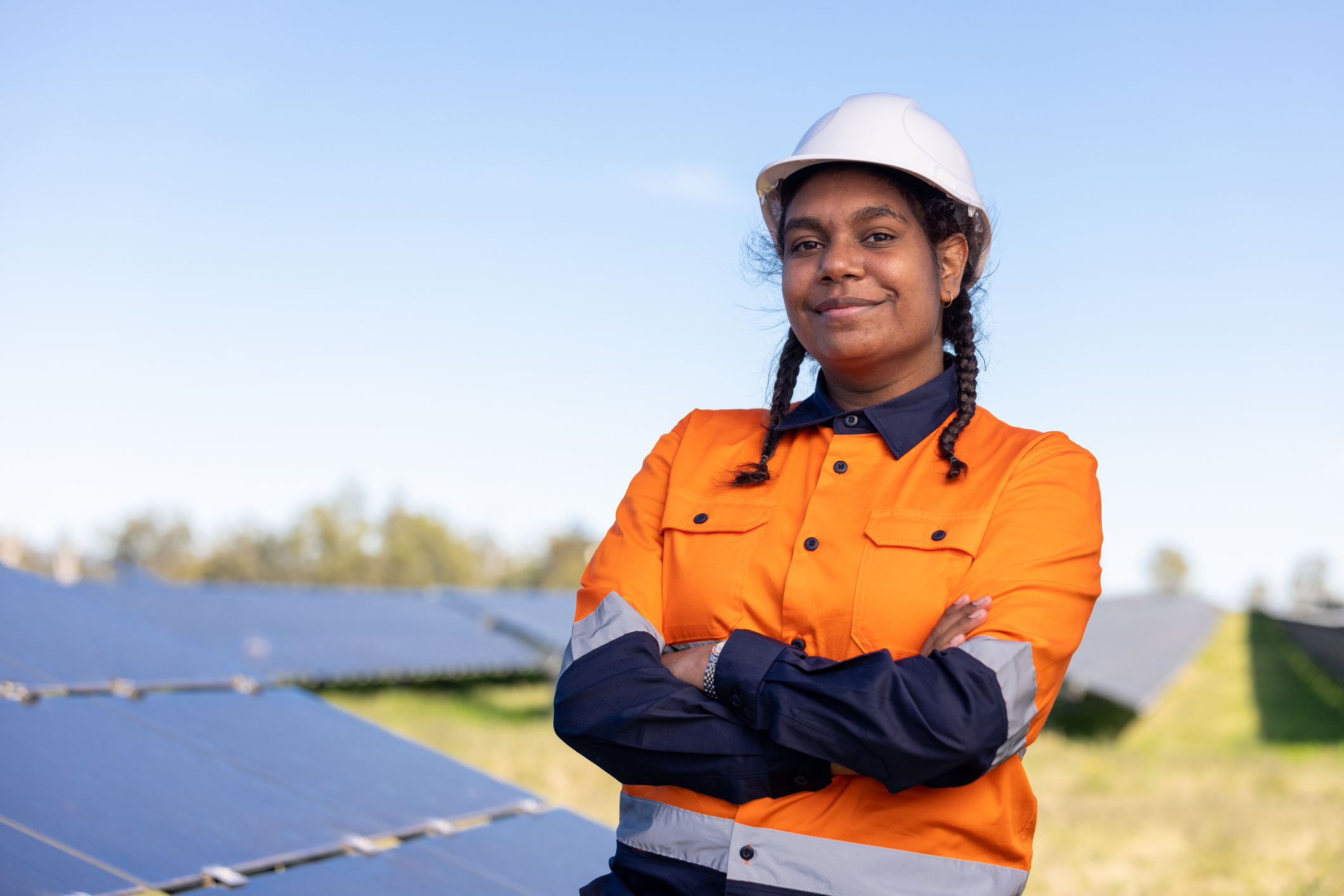 A young woman with brown skin and pigtail braids, wearing a hard hat and orange jacket, stands in front of solar panels.