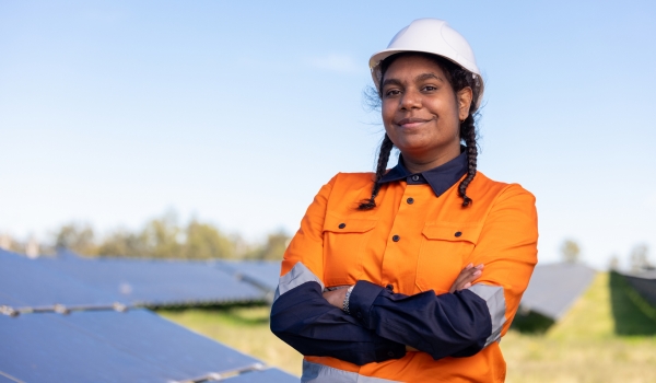 A young woman with brown skin and pigtail braids, wearing a hard hat and orange jacket, stands in front of solar panels.