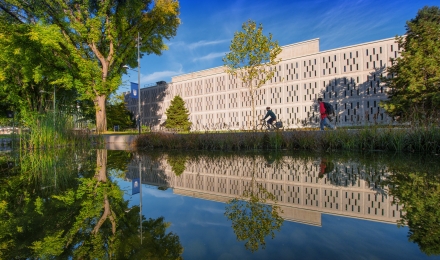 Two people on bikes pass between a pond surrounded by trees and greenery, and a university building, on the UBC campus.