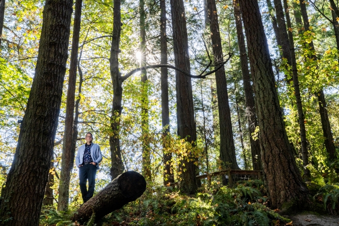 Dr. Ian Mauro, in a light blue jacket and dark blue printed shirt, stands on a log among the trees at Mystic Vale.