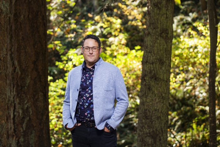 Dr. Ian Mauro, in a light blue jacket and dark blue printed shirt, stands among the trees at Mystic Vale.