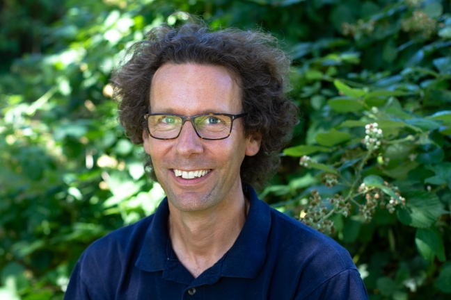 An image of Dr. Martin Scherwath smiling at the camera, wearing a dark blue shirt surrounded by trees and foliage. 
