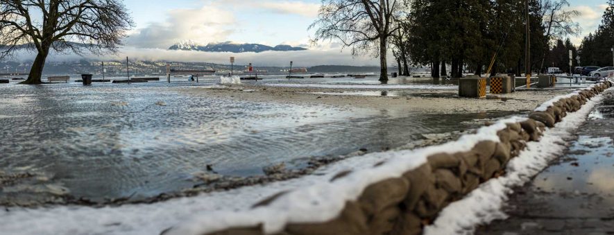 Jericho Beach Park in Vancouver during an episode of winter flooding. There are sandbags in the foreground, topped with snow. In mid-ground, the park grass is covered with a layer of water. The sea is beyond.