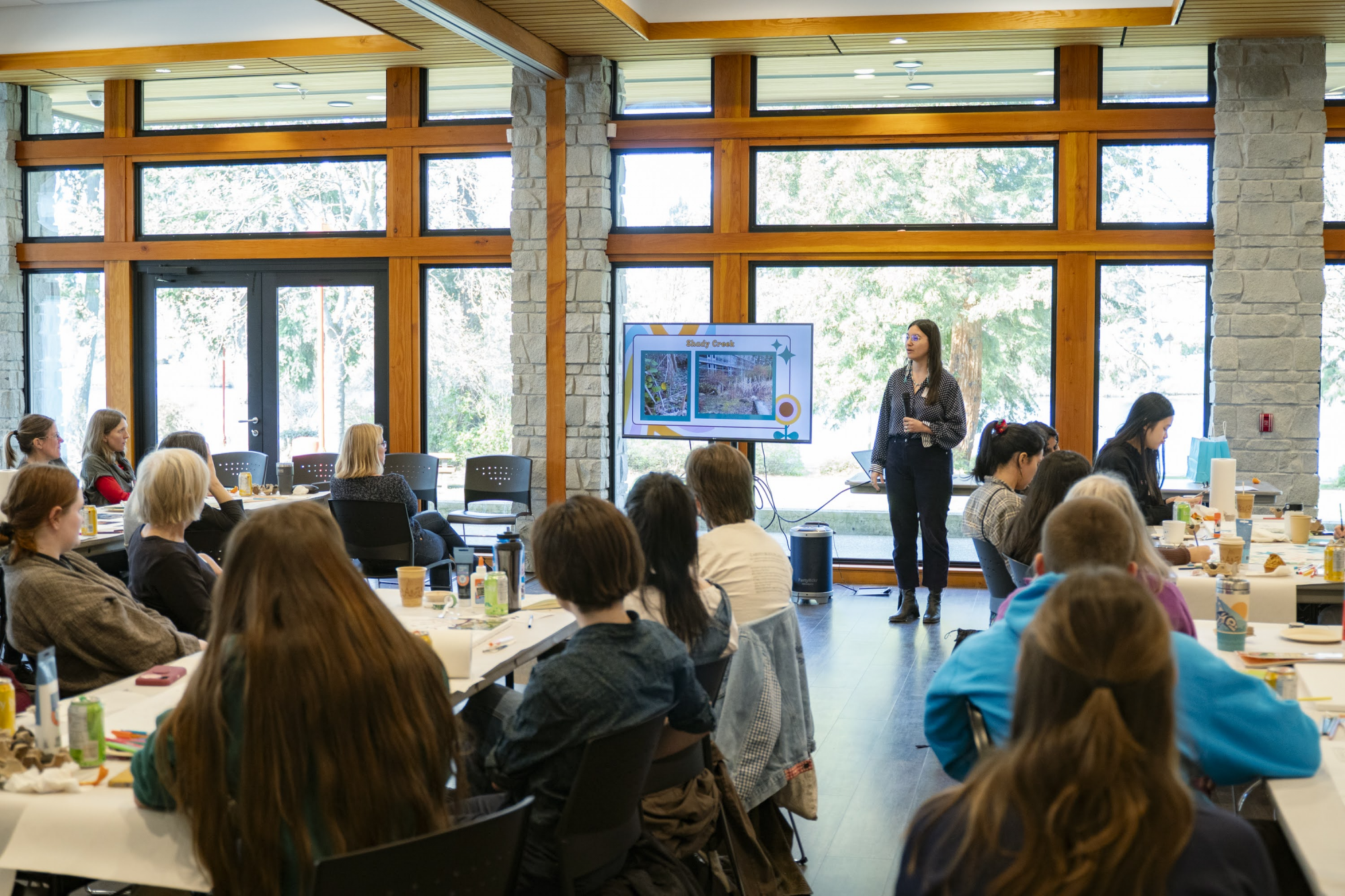 A person stands at the front of a room, giving a presentation to a crowd. Behind her, windows show trees.