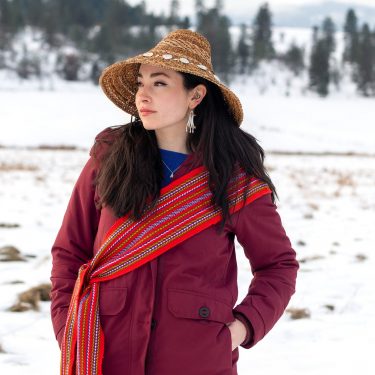 Janna Wale stands in front of a snow-swept landscape, wearing a Gitxsan cedar hat and a Métis sash.