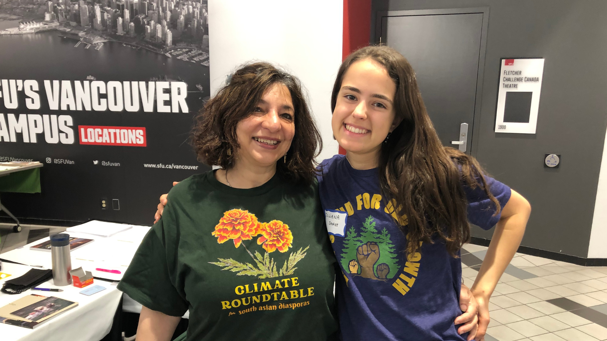 Two women smile for the camera with their arms around each other. They are inside a building with a sign that says "SFU's Vancouver Campus" behind them.